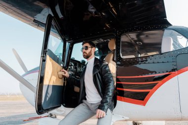 Sakallı, güneş gözlüklü, deri ceketli, helikoptere yaslanmış bir pilot. 