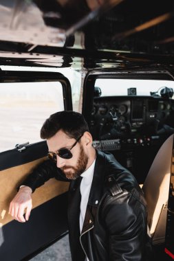 Şık güneş gözlüklü ve siyah deri ceketli sakallı pilot helikopter kapısının yanında.