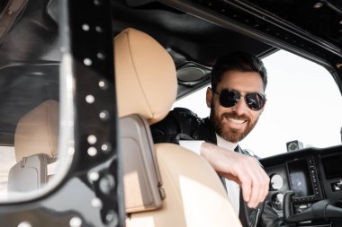 Havalı güneş gözlüklü ve deri ceketli pilot helikopter kokpitinde oturuyor. 