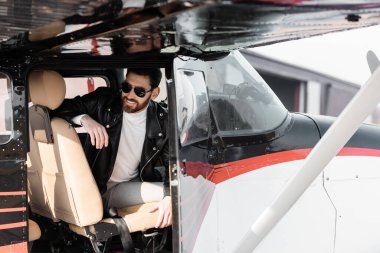 Havalı güneş gözlüklü ve deri ceketli sakallı pilot helikopter kokpitinde otururken gülümsüyor. 