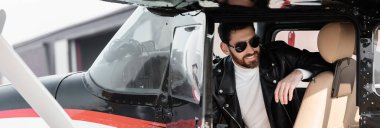 Havalı güneş gözlüklü ve deri ceketli sakallı pilot helikopter kokpitinde otururken gülümsüyor.