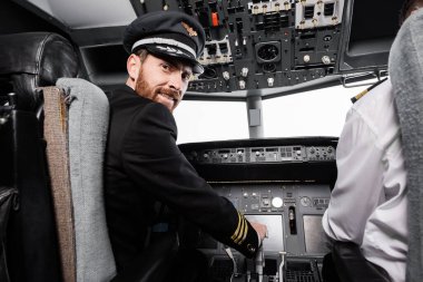 bearded pilot in cap smiling and looking at camera near co-pilot in airplane simulator  clipart