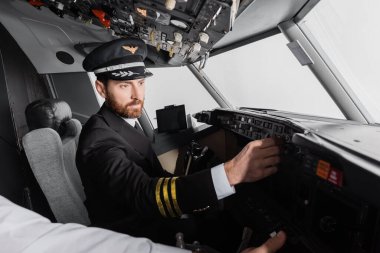 pilot in cap and uniform reaching control panel near co-pilot in airplane simulator clipart