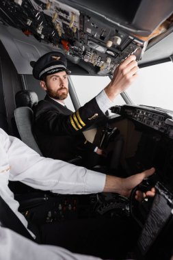 pilot in cap and uniform reaching overhead panel near co-pilot using yoke in airplane simulator clipart