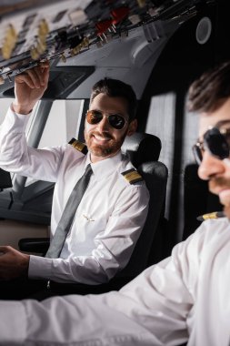 Güneş gözlüklü neşeli pilot uçak simülatöründe yardımcı pilot paneline yaklaşıyor.