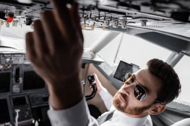 Güneş gözlüklü sakallı pilotun, uçak simülatöründe üst panele ulaşırken yüksek açılı görüntüsü