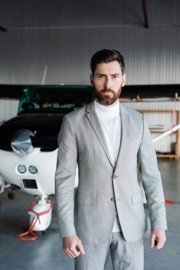 Gri takım elbiseli sakallı adam modern helikopterin yanında kameraya bakıyor. 