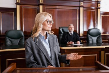  blonde advocate pointing with hand while talking in court near judge on background clipart