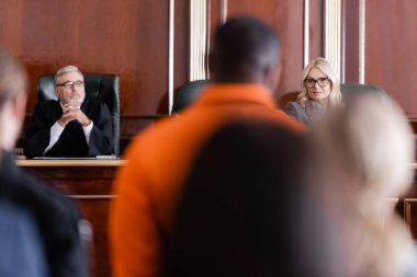Kıdemli yargıç elleri sımsıkı oturarak avukatın yanında oturuyor ve Afro-Amerikan bir adamı bulanık ön planda suçladı.