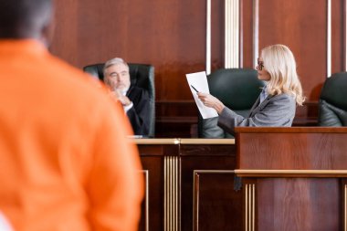Kıdemli yargıcın yanındaki davaya işaret eden ve Afro-Amerikan bir adamı bulanık ön planda suçlayan avukat.