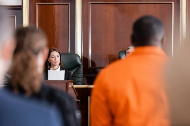 Savcı, yargıcın yanında dava okuyor ve bulanık önplanda Afro-Amerikan bir adamı suçluyor.