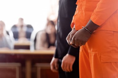 partial view of handcuffed african american man and bailiff near blurred jurors in court clipart
