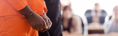 partial view of handcuffed african american man near blurred jurors in courtroom, banner clipart