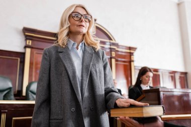 woman in eyeglasses giving swear on bible in court near prosecutor on blurred background clipart