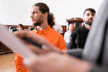 accused man looking away near guard and blurred prosecutor in courtroom clipart