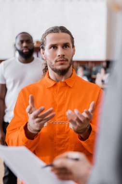 accused man in jail uniform and handcuffs gesturing near blurred prosecutor and african american juror in court clipart