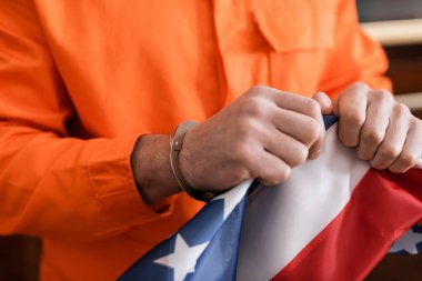 partial view of convicted man in handcuffs and jail uniform tearing usa flag, justice concept clipart