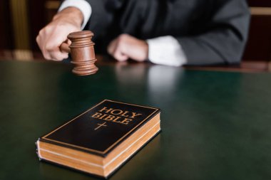 partial view of blurred judge holding gavel near holy bible on desk clipart