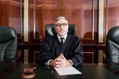 senior judge in mantle and eyeglasses sitting with clenched hands near gavel and paper on desk clipart