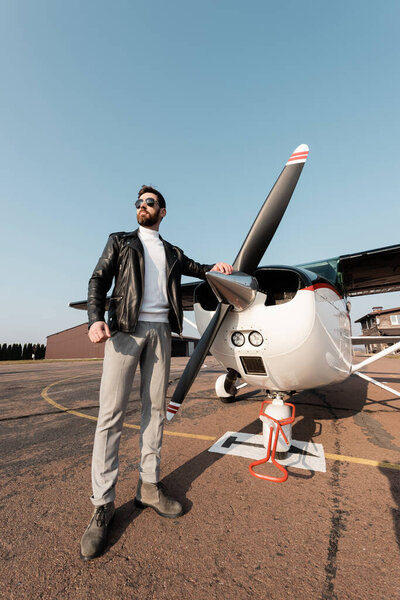 full length of bearded pilot in leather jacket and sunglasses standing near aircraft 