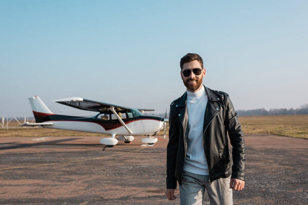 happy pilot in stylish leather jacket and sunglasses smiling near blurred helicopter