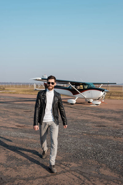 full length of positive pilot in stylish leather jacket and sunglasses walking near helicopter