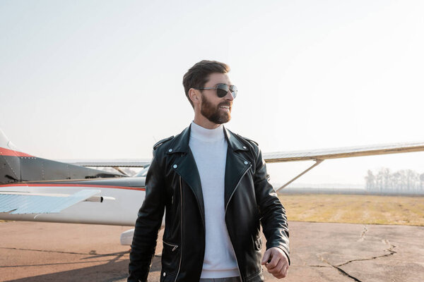 joyful pilot in stylish leather jacket and sunglasses near helicopter