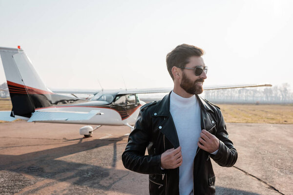 happy pilot in sunglasses adjusting stylish leather jacket  near helicopter