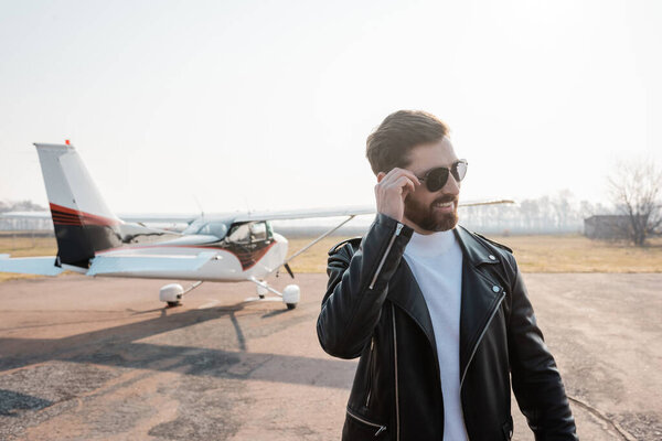 довольный пилот в стильной кожаной куртке регулировки солнцезащитных очков рядом с вертолетом