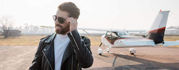 pleased pilot in stylish leather jacket adjusting sunglasses near helicopter, banner