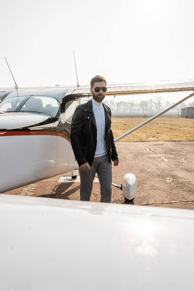 bearded pilot in black leather jacket and sunglasses standing near helicopter