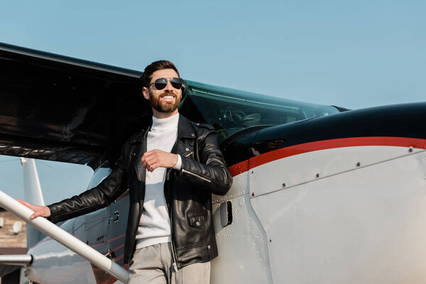 счастливый бородатый мужчина в солнцезащитных очках и черной кожаной куртке стоит рядом с вертолетом