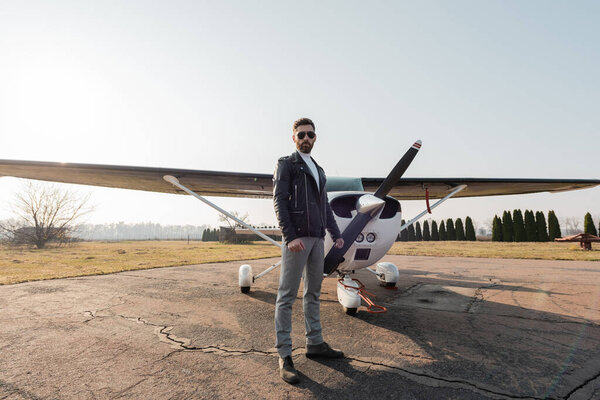 полная длина бородатого пилота в стильных солнцезащитных очках и кожаной куртке, стоящих рядом с современным самолетом 