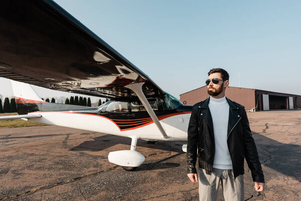 бородатый мужчина в стильных солнцезащитных очках и кожаной куртке стоит рядом с современным самолетом 