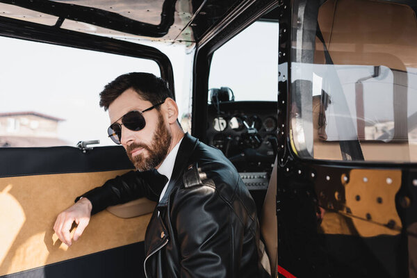 bearded pilot in stylish sunglasses and leather jacket near opened door of helicopter 