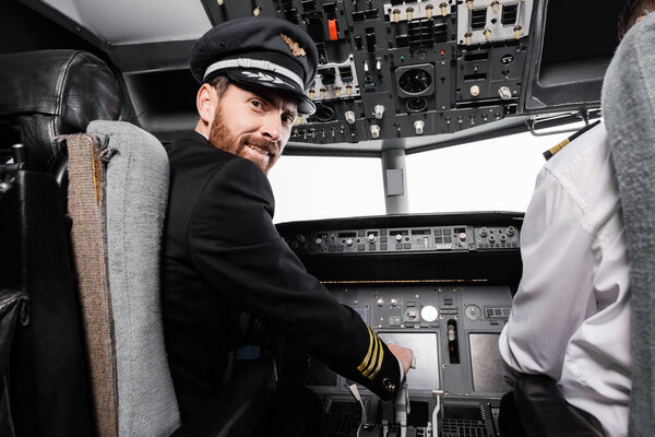 bearded pilot in cap smiling and looking at camera near co-pilot in airplane simulator 