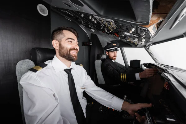 cheerful co-pilot using yoke near captain reaching control panel in airplane simulator