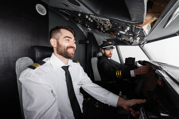 cheerful co-pilot using yoke near captain reaching control panel in airplane simulator 