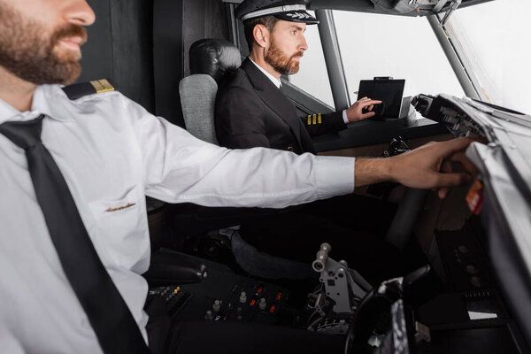 captain in cap and uniform using device with blank screen near co-pilot in airplane simulator