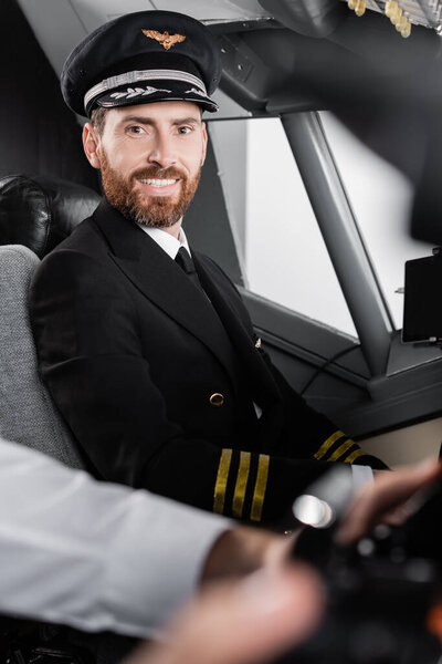 bearded pilot in uniform and cap smiling near blurred co-pilot 