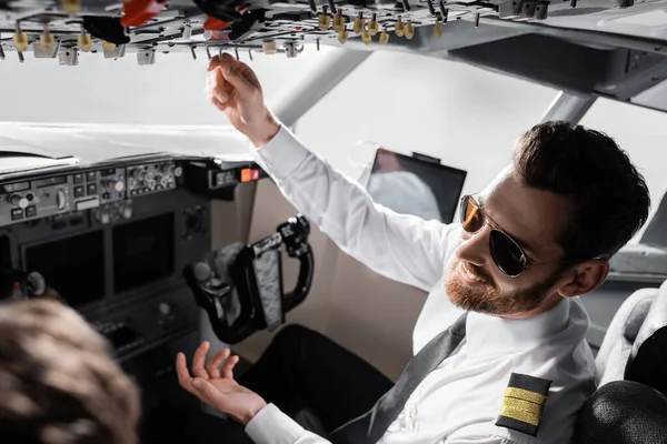 cheerful pilot in sunglasses reaching overhead panel near co-pilot in plane simulator