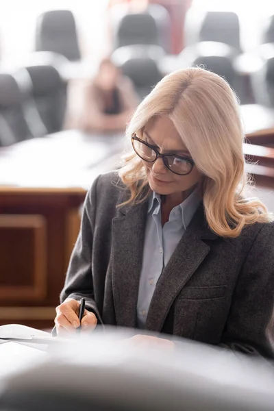 blonde middle aged advocate in courtroom writing on blurred foreground