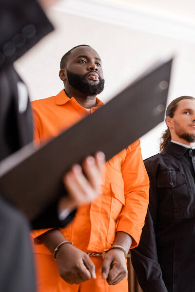 accused african american man in handcuffs standing near bailiff and blurred prosecutor with clipboard in court