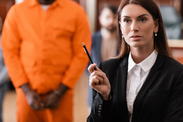 brunette prosecutor in formal wear standing with pen near accused african american man on blurred background