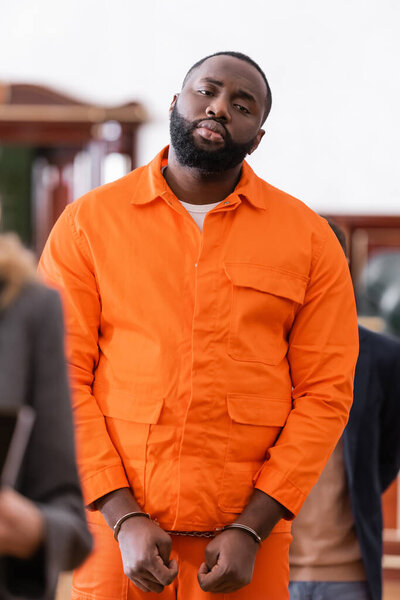 handcuffed african american man in orange jail uniform in courtroom on blurred foreground