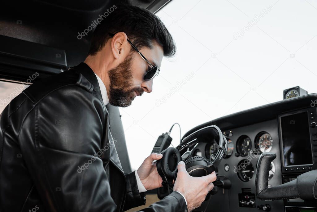 bearded pilot in sunglasses and black leather jacket holding headphones in cockpit of helicopter 