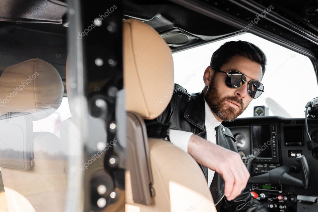 bearded pilot in stylish sunglasses and leather jacket sitting in cockpit of helicopter 