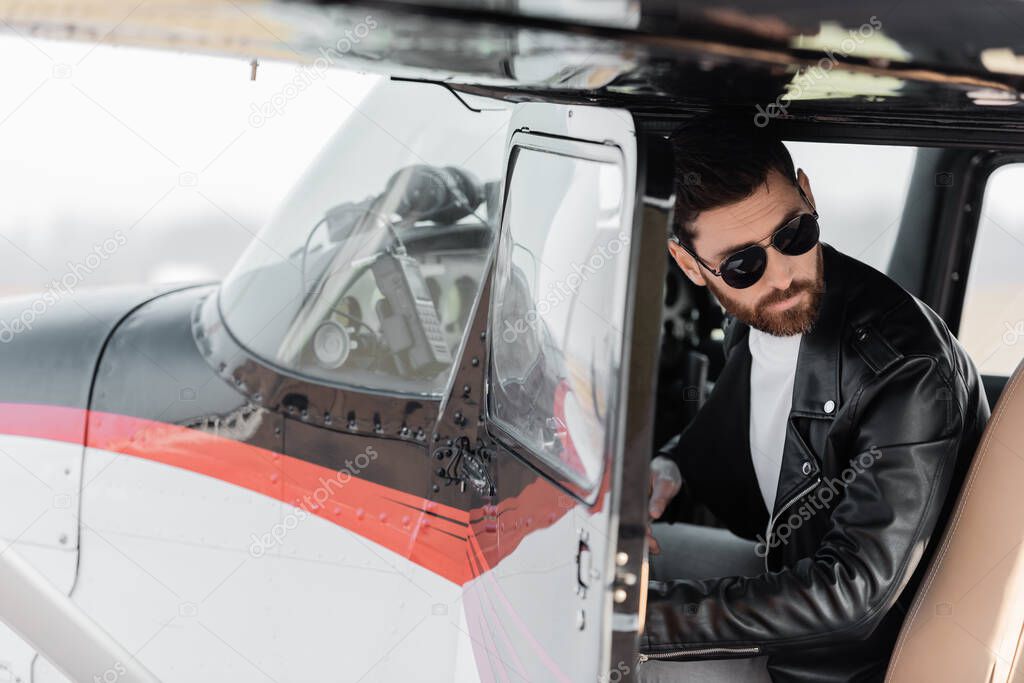 bearded pilot in sunglasses and leather jacket opening door of helicopter and looking away 