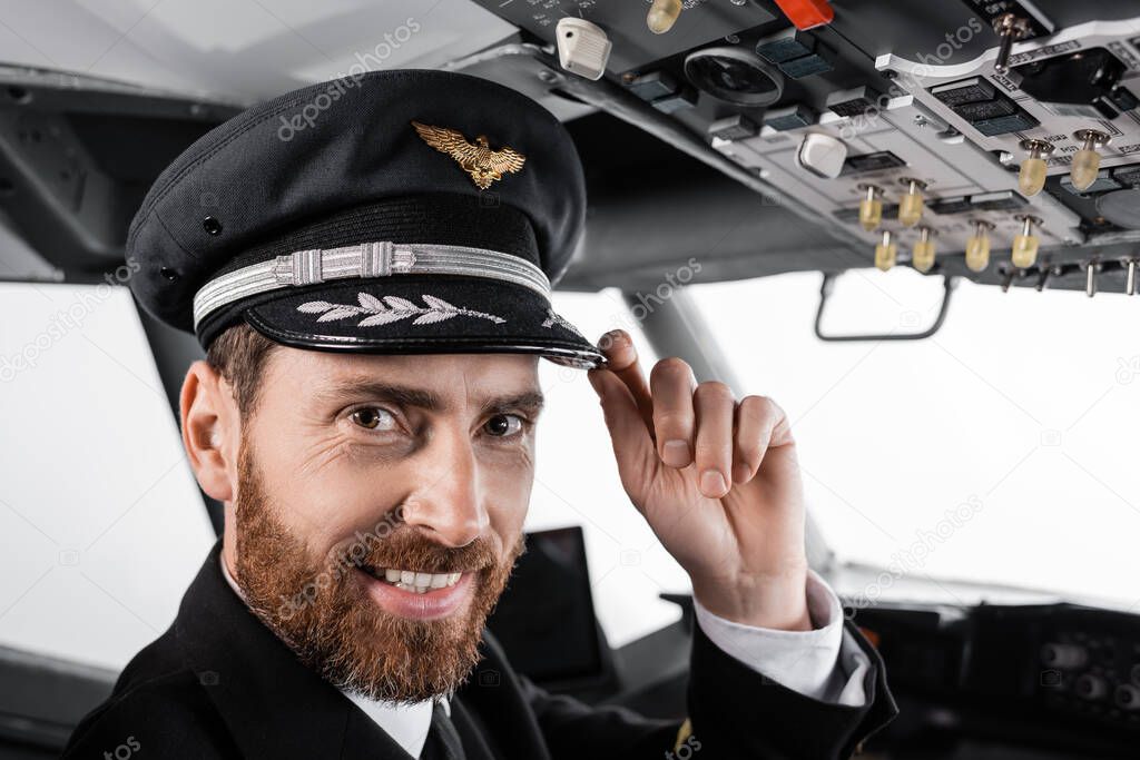 cheerful pilot adjusting cap and looking at camera in airplane simulator 