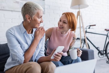Gülümseyen lezbiyen kadınlar bir fincan çay ve akıllı telefonla evdeki kanepede konuşuyorlar.